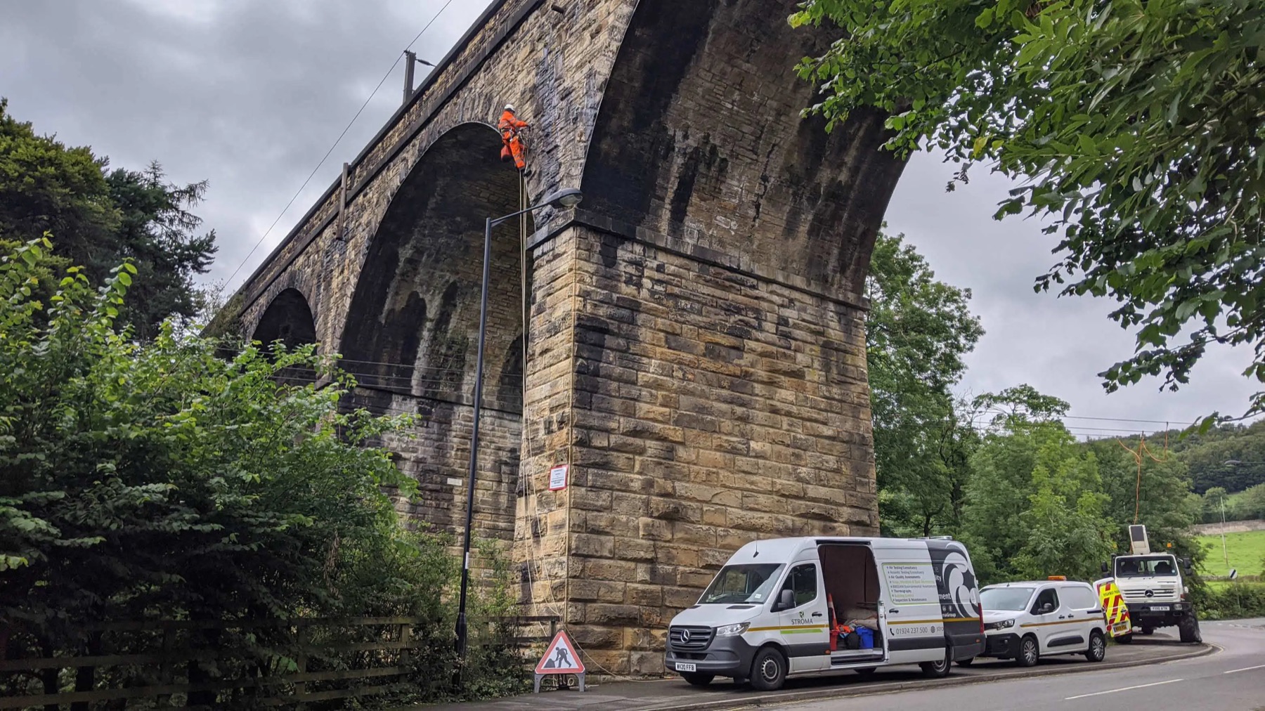 A Stroma rope access technician suspended from a stone railway viaduct, with the Stroma van and traffic management on the road below